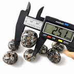 Digital caliper measuring a VoraZen™ Yooperlite stone, showing a size between 25mm and 28mm. The professional measurement ensures accuracy for these fluorescent sodalite-rich syenite tumbled stones, perfect for UV light displays.