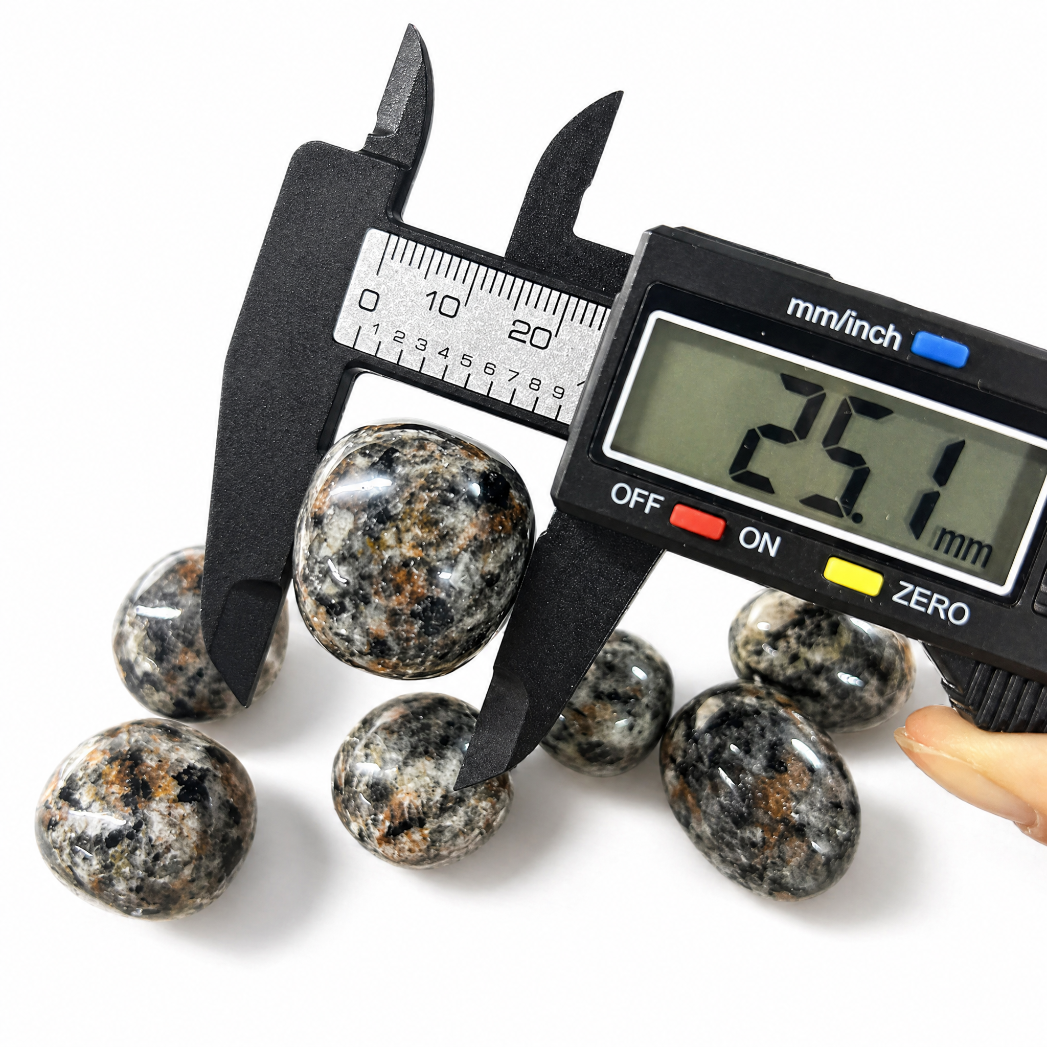 Digital caliper measuring a VoraZen™ Yooperlite stone, showing a size between 25mm and 28mm. The professional measurement ensures accuracy for these fluorescent sodalite-rich syenite tumbled stones, perfect for UV light displays.