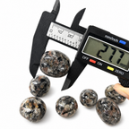 Digital caliper measuring a VoraZen™ Yooperlite stone, showing a size between 25mm and 28mm. The professional measurement ensures accuracy for these fluorescent sodalite-rich syenite tumbled stones, perfect for UV light displays.